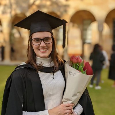 A young woman faces the camera wearing graduation robe and cap, smiling. She is holding a bunch of flowers.